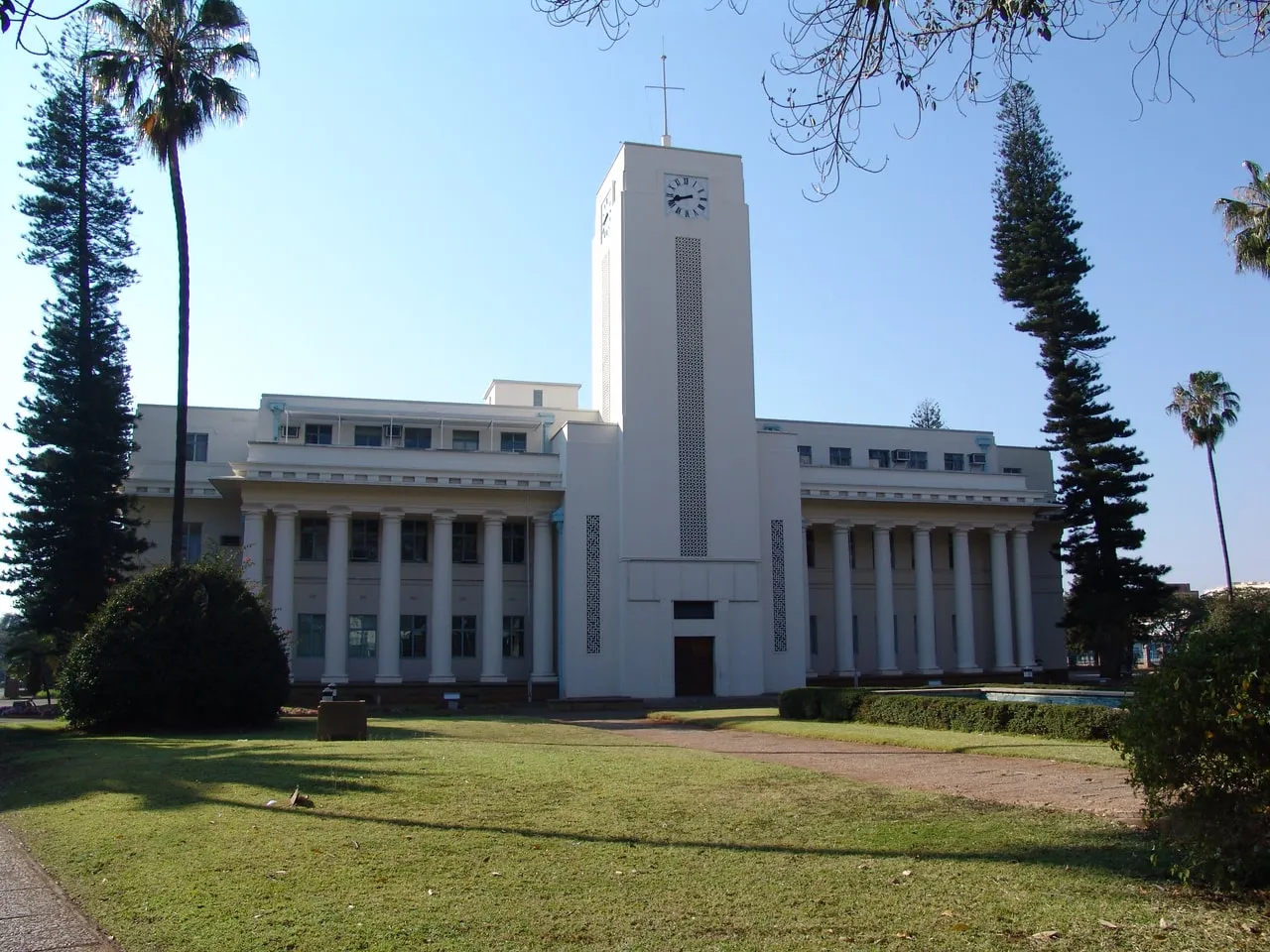 Bulawayo City Hall head-on across the lawn: a long colonnade either side of a central clock tower, with pines and palms framing it.